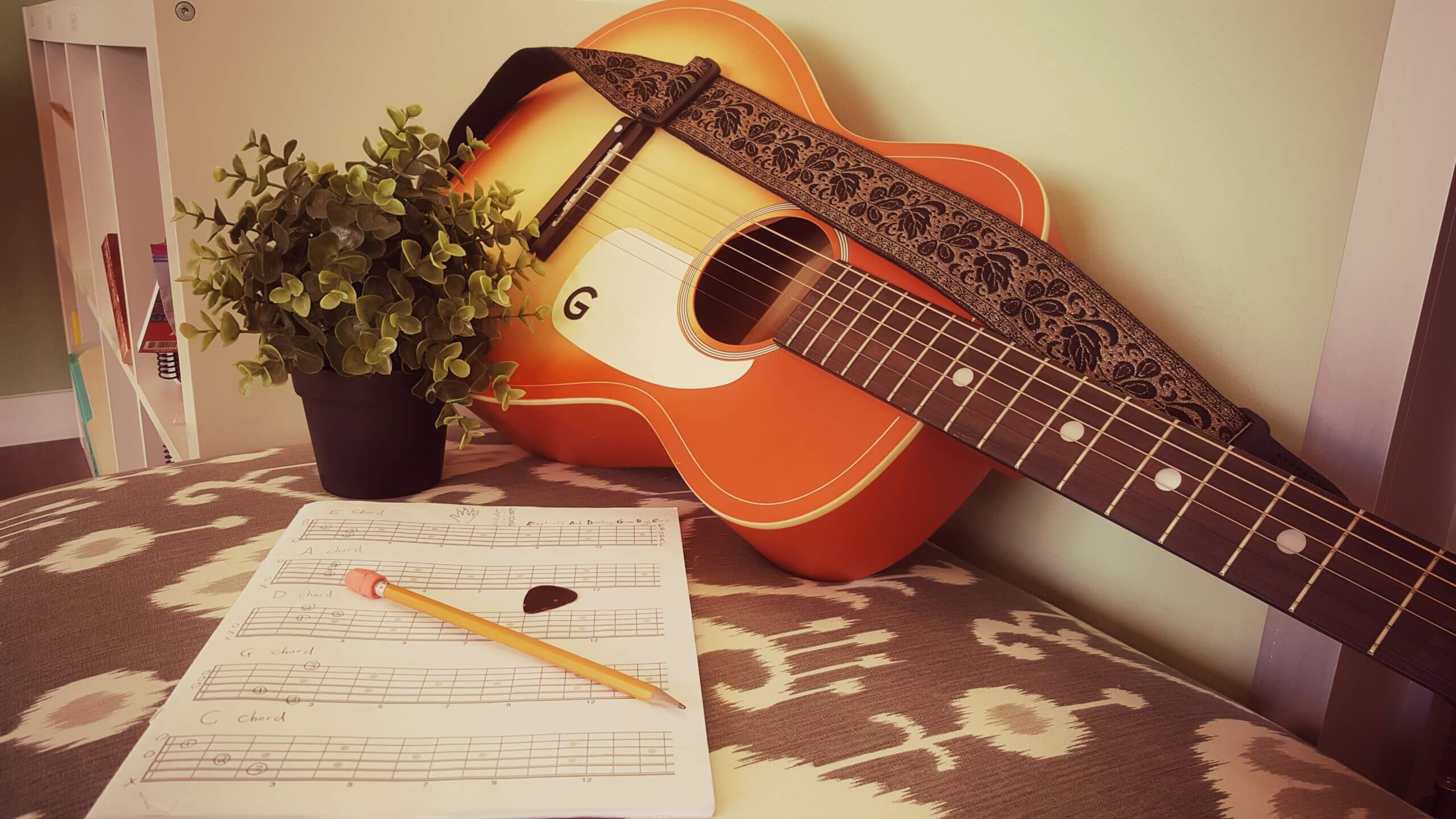 An acoustic guitar with a strap leans against a wall next to a small potted plant. On the table in the foreground, there is sheet music, a pencil, and a guitar pick.