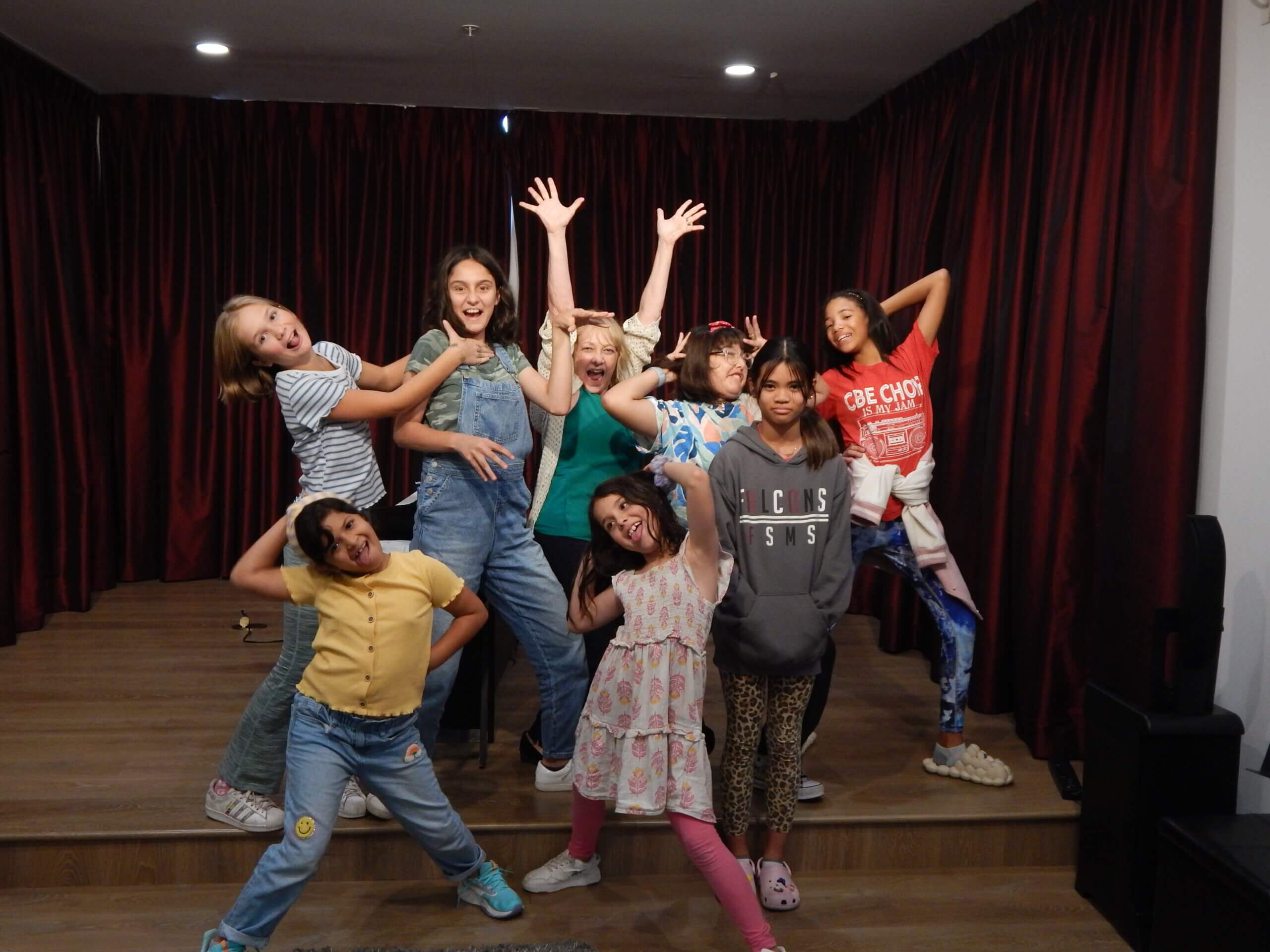 Acting class. Kids and teacher are on the stage with hands put up.